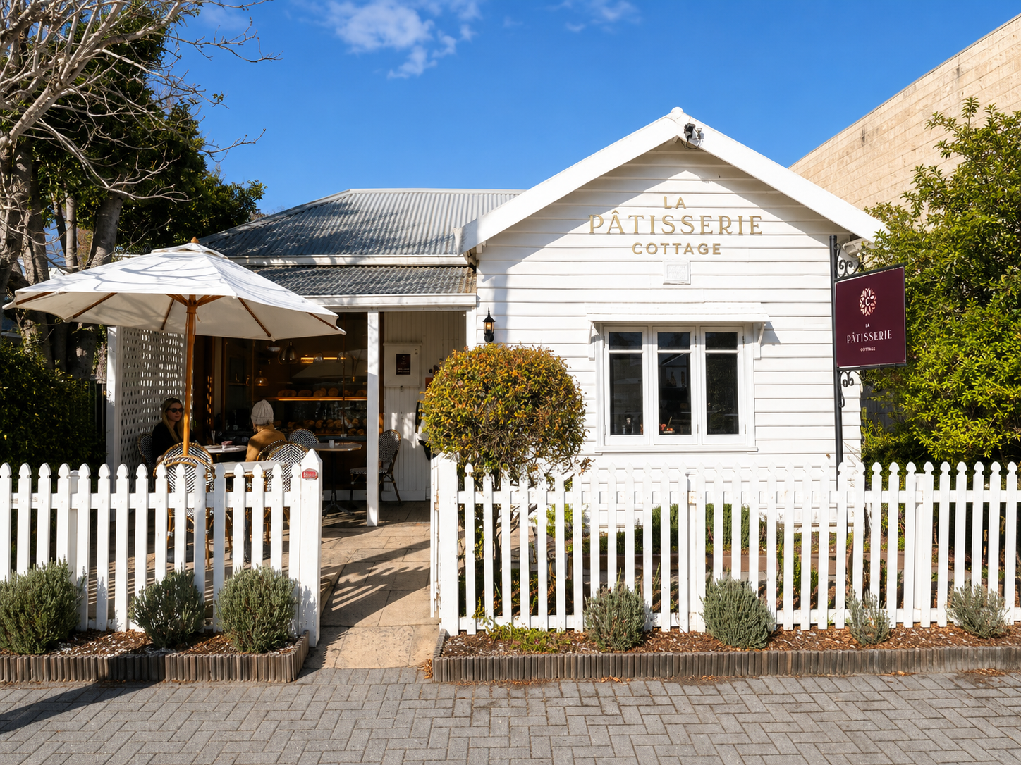 The white weatherboard cottage of La Pâtisserie Cottage on Peels Place, with a parasol, picket fence and lavender garden under a blue sky